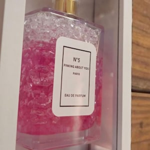 K&ouml;nnte beinhalten: Ein Parf&uuml;mflakon mit goldenem Verschluss und einem transparenten, facettierten Deckel wird in einer wei&szlig;en Schattenbox pr&auml;sentiert. Die Flasche ist mit rosa Fl&uuml;ssigkeit gef&uuml;llt und tr&auml;gt das Etikett "N&deg;5 PINKING ABOUT YOU PARIS EAU DE PARFUM."