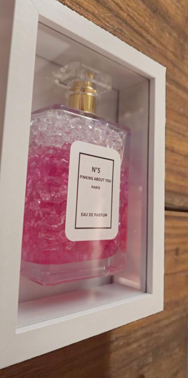 K&ouml;nnte beinhalten: Ein Parf&uuml;mflakon mit goldenem Verschluss und einem transparenten, facettierten Deckel wird in einer wei&szlig;en Schattenbox pr&auml;sentiert. Die Flasche ist mit rosa Fl&uuml;ssigkeit gef&uuml;llt und tr&auml;gt das Etikett "N&deg;5 PINKING ABOUT YOU PARIS EAU DE PARFUM."