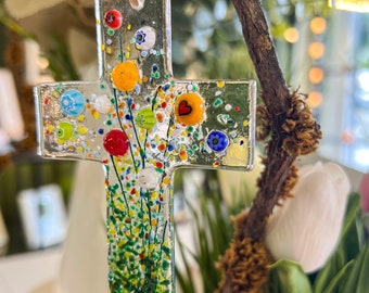 May include: A clear glass cross ornament with colorful floral accents. The cross features red, orange, blue, and green glass beads, and is suspended by a natural twine loop. The design includes a heart-shaped detail.