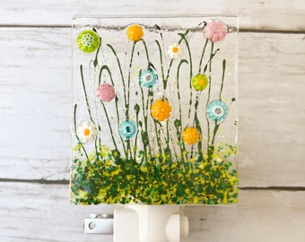 May include: A decorative night light featuring a clear glass panel with a colorful floral design. The design includes green stems, yellow and green foliage, and various colored flowers, including pink, orange, and white. The night light has a white plug.