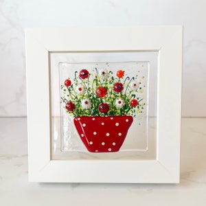 May include: A framed glass art piece featuring a red polka-dotted flower pot filled with red and white flowers. The artwork is encased in a white frame, creating a visually appealing display. The flowers have red and white petals with green stems.