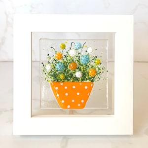 May include: A framed glass art piece featuring a colorful floral arrangement in an orange polka-dotted pot. The flowers are in shades of blue, orange, white, and green. The artwork is set against a clear background and framed in a white square frame.