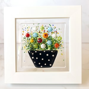 May include: A framed glass art piece featuring a colorful floral arrangement. The flowers are in a black polka-dotted vase. The flowers are various colors, including red, orange, blue, and white. The frame is white.