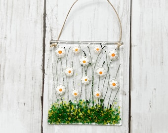 May include: A rectangular glass wall hanging featuring a field of white daisies with orange centers and green stems. The daisies are set against a clear background, with a brown twine loop for hanging. The bottom has green and yellow textured glass.