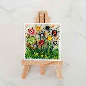 May include: A square fused glass art piece featuring a colorful floral meadow design with various millefiori glass flowers in red, yellow, green, and black, displayed on a small wooden easel. The glass panel has a clear base with green foliage.