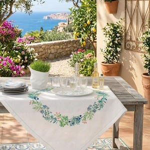 Peut inclure: Une nappe blanche avec une bordure florale est posée sur une table en bois. La table est dressée avec des assiettes, des verres et des bouteilles. L'arrière-plan présente une vue panoramique sur la mer et la verdure.