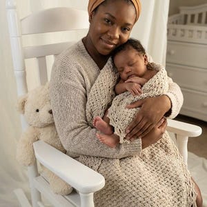 Puede incluir: Una mujer con un suéter beige y un pañuelo en la cabeza acuna a un bebé dormido envuelto en una manta de crochet a juego. Una mecedora blanca y un oso de peluche también están en la imagen.