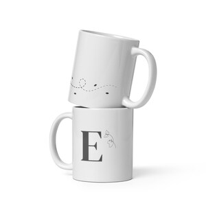 May include: Two white ceramic mugs stacked on top of each other. The top mug has a dotted line design with small black dots. The bottom mug features the letter "E" in a dark gray color, with a small floral design.
