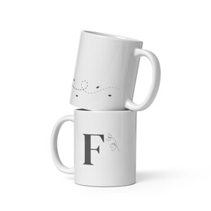 Può includere: Due tazze in ceramica bianca impilate l'una sull'altra. La tazza superiore presenta un semplice disegno di una linea punteggiata con un anello all'inizio. La tazza inferiore ha una lettera "F" nera e un piccolo disegno floreale.