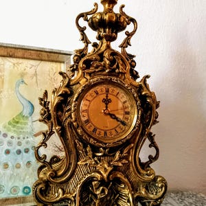 May include: A gold-toned, ornate, decorative clock with a Roman numeral clock face. The clock has a detailed, intricate design with a floral motif.
