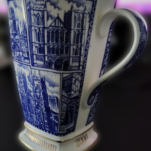 May include: A white ceramic mug featuring blue illustrations of cathedrals, including Ripon Cathedral. The base is inscribed with the gold text Millennium 2000. The mug has a curved handle and a gold-trimmed rim and base, showcasing detailed architectural sketches.