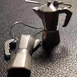 May include: A pair of silver-coloured coffee pot earrings. Each earring is a miniature replica of a classic stovetop espresso maker, complete with a handle and spout. The earrings are suspended from a simple hook.