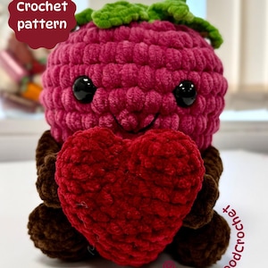 Peut inclure: Peluche de framboise au crochet tenant un cœur rouge. La framboise est rose avec des feuilles vertes et des yeux noirs. Le texte "Crochet pattern" est dans un cercle rouge. Le texte "@WheelyGoodCrochet" est également visible.