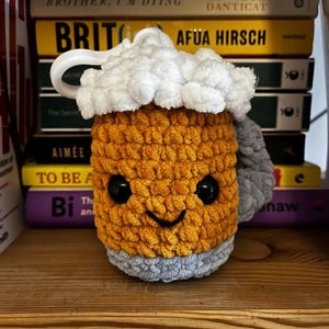 May include: A crocheted beer mug plush toy with a white foam top, brown beer, and a gray handle. The mug has a smiling face with black eyes.