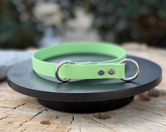 May include: A lime green collar featuring a smooth, flexible strap material. The collar includes two silver-toned metal D-rings positioned on either side of a central overlapping section, which is secured by two small, circular silver-toned metal rivets.