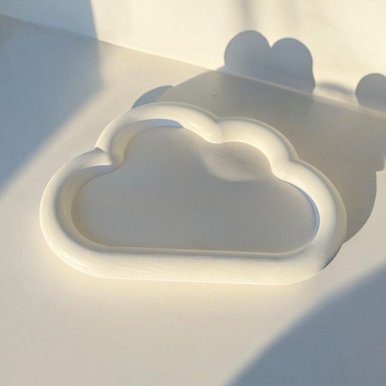 May include: A white, cloud-shaped tray with a raised rim. The tray is made of a solid material and sits on a white surface. The image is lit with natural light, casting shadows.
