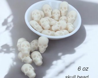 May include: A white ceramic bowl filled with small, off-white skull-shaped objects. Some skulls are scattered outside the bowl. The text "6 oz skull head" is visible in the image. The objects appear to be wax melts or decorative items.