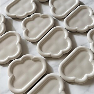 May include: A collection of off-white, cloud-shaped trays. Each tray has a rounded, raised edge and a smooth, flat interior. The trays are arranged on a white marble surface, creating a minimalist aesthetic. These trays are ideal for holding small items.