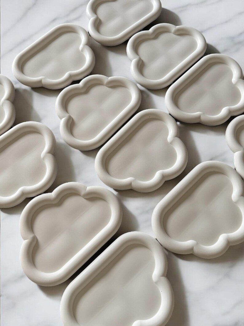 May include: A collection of off-white, cloud-shaped trays. Each tray has a rounded, raised edge and a smooth, flat interior. The trays are arranged on a white marble surface, creating a minimalist aesthetic. These trays are ideal for holding small items.