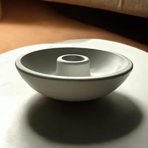 May include: A light gray, modern candle holder with a circular bowl shape and a central, cylindrical pillar. The holder is made of a smooth, matte material and casts a dark shadow on the surface below.