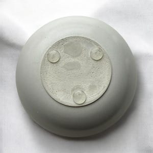 May include: A light gray, round concrete dish with a textured base. Three clear, rounded feet are attached to the base. The dish is sitting on a white surface.