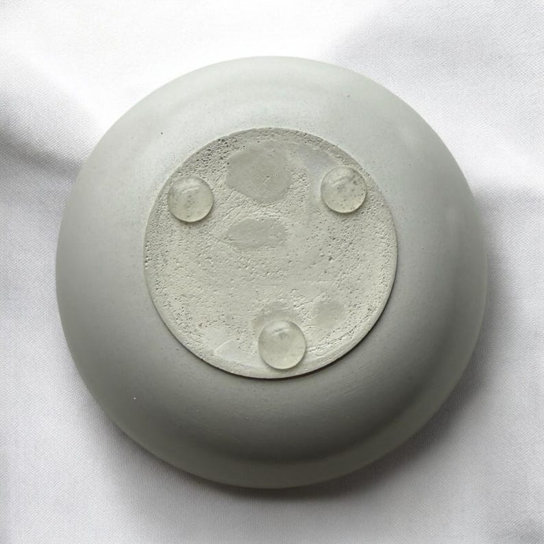 May include: A light gray, round concrete dish with a textured base. Three clear, rounded feet are attached to the base. The dish is sitting on a white surface.