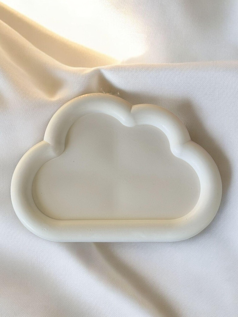 May include: A white cloud-shaped tray made of a solid material. The tray has a smooth, rounded edge and a shallow interior. The background is a white fabric with soft folds.