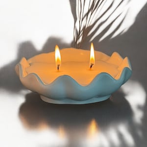 May include: A lit, two-wick candle in a light blue, shell-shaped ceramic container. The candle wax is a creamy white, and the flames are bright yellow-orange. The background is a soft white with shadows.