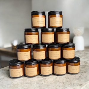 Puede incluir: Una pirámide de doce velas perfumadas en frascos de vidrio con tapas negras. Cada vela tiene una etiqueta con un nombre de aroma diferente, como "Salted Caramel" y "Lavender". Las velas están dispuestas sobre una encimera clara.