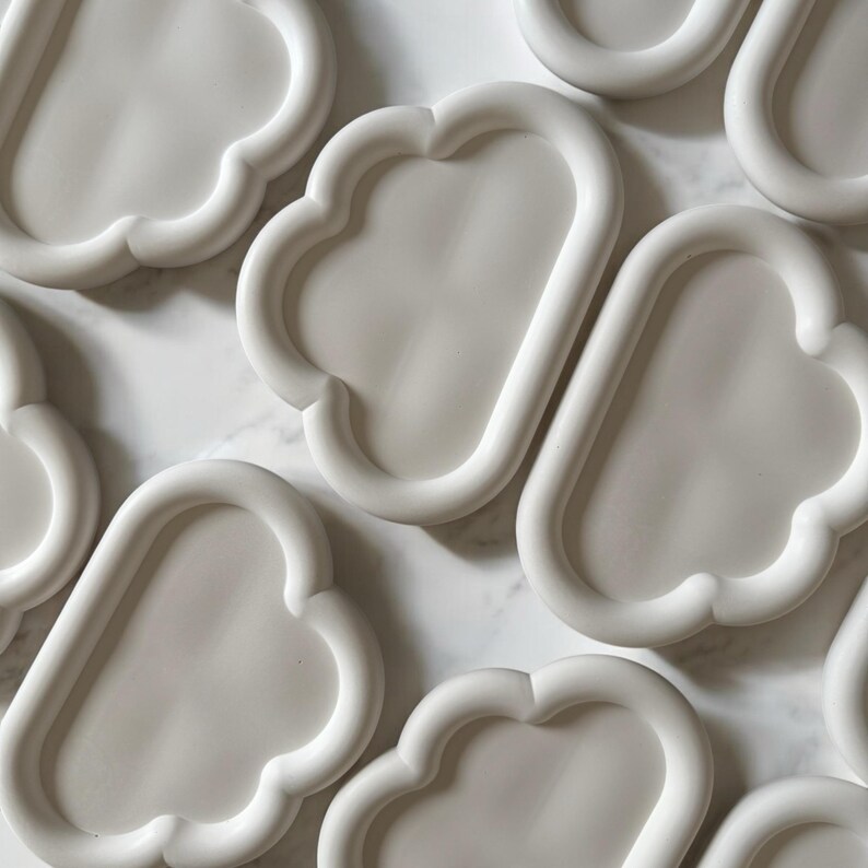 May include: A collection of white, cloud-shaped trays. Each tray has a rounded, oblong interior with a raised, scalloped edge. The trays are arranged on a light-colored, possibly marble, surface. The overall aesthetic is minimalist and modern.