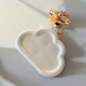 May include: A white cloud-shaped tray with a raised edge. A gold-colored chain with a pendant and a small ring are on the tray. The pendant has a woven design and orange accents. The tray is on a light gray surface.