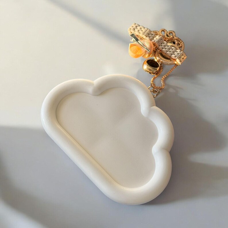 May include: A white cloud-shaped tray with a raised edge. A gold-colored chain with a pendant and a small ring are on the tray. The pendant has a woven design and orange accents. The tray is on a light gray surface.