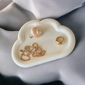 May include: A white cloud-shaped trinket tray holding a gold necklace, a gold ring, and a seashell ring. The tray is on a gray surface. The jewelry tray is a decorative item for storing small accessories.