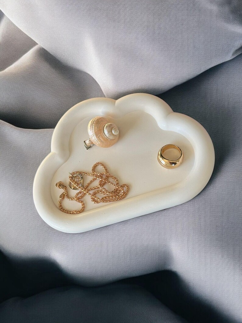 May include: A white cloud-shaped trinket tray holding a gold necklace, a gold ring, and a seashell ring. The tray is on a gray surface. The jewelry tray is a decorative item for storing small accessories.