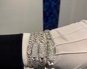 Peut inclure: Quatre bracelets argentés incrustés de diamants. Les bracelets présentent un design à maillons gourmette, chaque maillon étant recouvert de petits diamants étincelants. Ils sont présentés sur une main gantée de blanc, avec une manche noire visible.