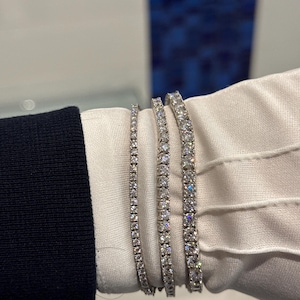 May include: Three diamond tennis bracelets of varying widths are displayed. The bracelets are set in a silver metal and feature multiple round-cut diamonds. The bracelets are shown against a white glove and a dark blue sleeve.