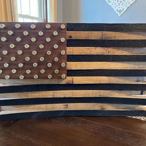 May include: A wooden American flag wall decor with a distressed finish. The flag is made of reclaimed wood and features a field of stars made of bullet casings.