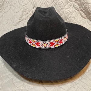 Puede incluir: Un sombrero vaquero de fieltro negro con una banda de cuentas. La banda es roja, blanca y amarilla con un patrón geométrico.