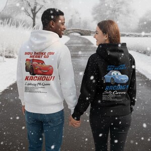 May include: Two people wearing matching hoodies, walking on a snowy path. One hoodie is white with a red Lightning McQueen graphic and the text "YOU MAKE MY HEART GO KACHOW!". The other is black with a blue Sally Carrera graphic and the text "YOU LIVE MY HEART RACING!".