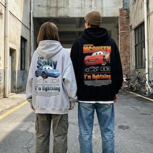 May include: Two people wearing matching hoodies featuring cartoon car designs. One is grey with a blue car and the text "I'm Sally" and "I'm lightning." The other is black with a red car and the text "Lightning McQueen" and "I'm lightning."