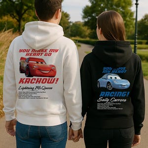 Op de afbeelding: Twee mensen dragen bijpassende hoodies. De witte hoodie heeft Lightning McQueen met "YOU MAKE MY HEART GO" en "KACHOW!". De zwarte hoodie toont Sally Carrera met "YOU HAVE MY HEART" en "RACING!" op de rug.