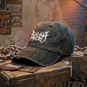 May include: A black denim baseball cap with white embroidered text. The cap sits on a wooden box with patches. The background includes concert posters and a metal chain.