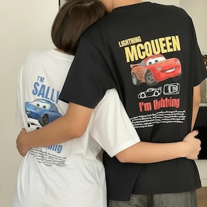 Könnte beinhalten: Schwarze und weiße T-Shirts mit Cartoon-Autografiken. Das schwarze Shirt zeigt Lightning McQueen mit Text, und das weiße Shirt hat Sally Carrera. Beide Shirts haben Text auf der Rückseite. Freizeitkleidung.