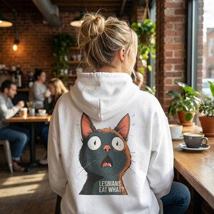 Könnte beinhalten: Weißer Kapuzenpullover mit einem überraschten Katzen-Design in Schwarz, Orange und Weiß. Die Katze hat einen erstaunten Gesichtsausdruck und der Text "LESBIANS EAT WHAT?" steht darunter. Der Hoodie hat eine Kapuze und eine Vordertasche.