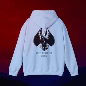 May include: A white hooded sweatshirt with a black dragon graphic on the back. The dragon has large wings and a detailed design. The text "KNEEL BEFORE THE ALPHA" is below the dragon. The background is a gradient of red, blue, and black.