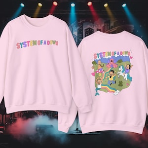 Puede incluir: Sudadera rosa claro con el texto "SYSTEM OF A DOWN" en letras multicolores. La parte trasera de la sudadera presenta una ilustración de dibujos animados con un castillo, un arcoíris y varios personajes.