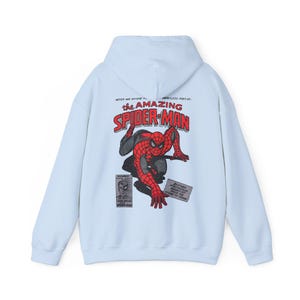Może przedstawiać: Jasnoniebieska bluza z kapturem z grafiką Spider-Mana w kolorze czerwonym i czarnym, z napisem "The Amazing Spider-Man" w kolorze czerwonym. Bluza ma kaptur ze sznurkiem i długie rękawy. Projekt komiksowy.