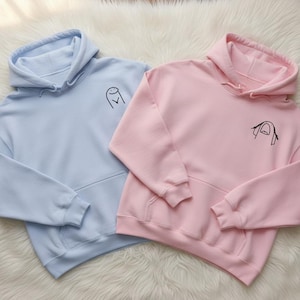 Könnte beinhalten: Zwei Kapuzenpullover, einer hellblau und einer rosa, werden gezeigt. Jeder hat eine Vordertasche und eine Kapuze mit Kordelzug. Auf der Brust jedes Sweatshirts befindet sich eine kleine schwarze Strichzeichnung. Die Sweatshirts liegen auf einer weißen, flauschigen Oberfläche.