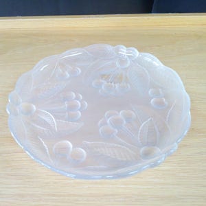Puede incluir: Un plato redondo de cristal transparente con un diseño en relieve de cerezas y hojas. El plato tiene un borde festoneado y probablemente se utiliza para servir o exhibir. El diseño está grabado en el cristal, creando una textura sutil y elegante.