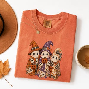 Pode incluir: Uma t-shirt de gola redonda em cor laranja queimado com um gráfico de três fantasmas usando chapéus de bruxa estampados e segurando abóboras. Os fantasmas estão decorados com várias estampas de outono, incluindo morcegos, flores e xadrez.